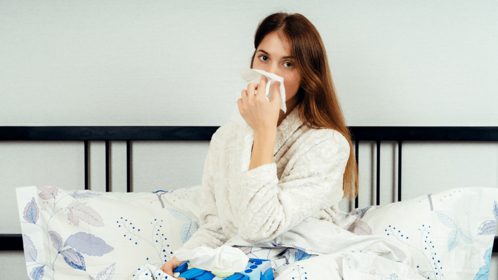 5 Tips for an Allergy-Proof Bedroom Feature Image: Young woman sitting alone on the bed with a napkin at her nose. Concept of flu or allergic disease.